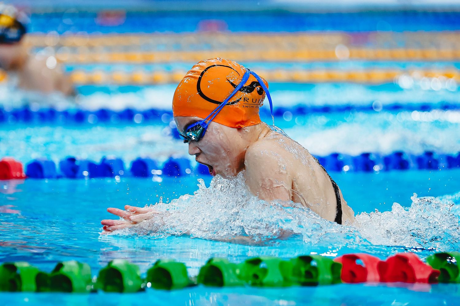 Female swimmer in orange swimsuit and blue cap stands poolside at an indoor swimming competition.