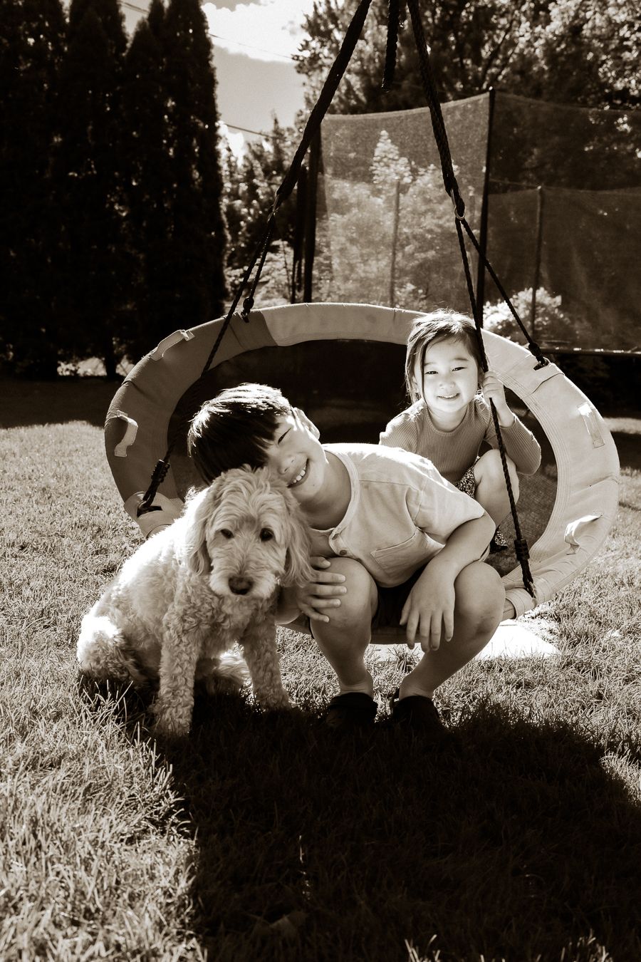 Two children and a fluffy golden doodle dog pose together in a backyard near a trampoline in sepia tone.