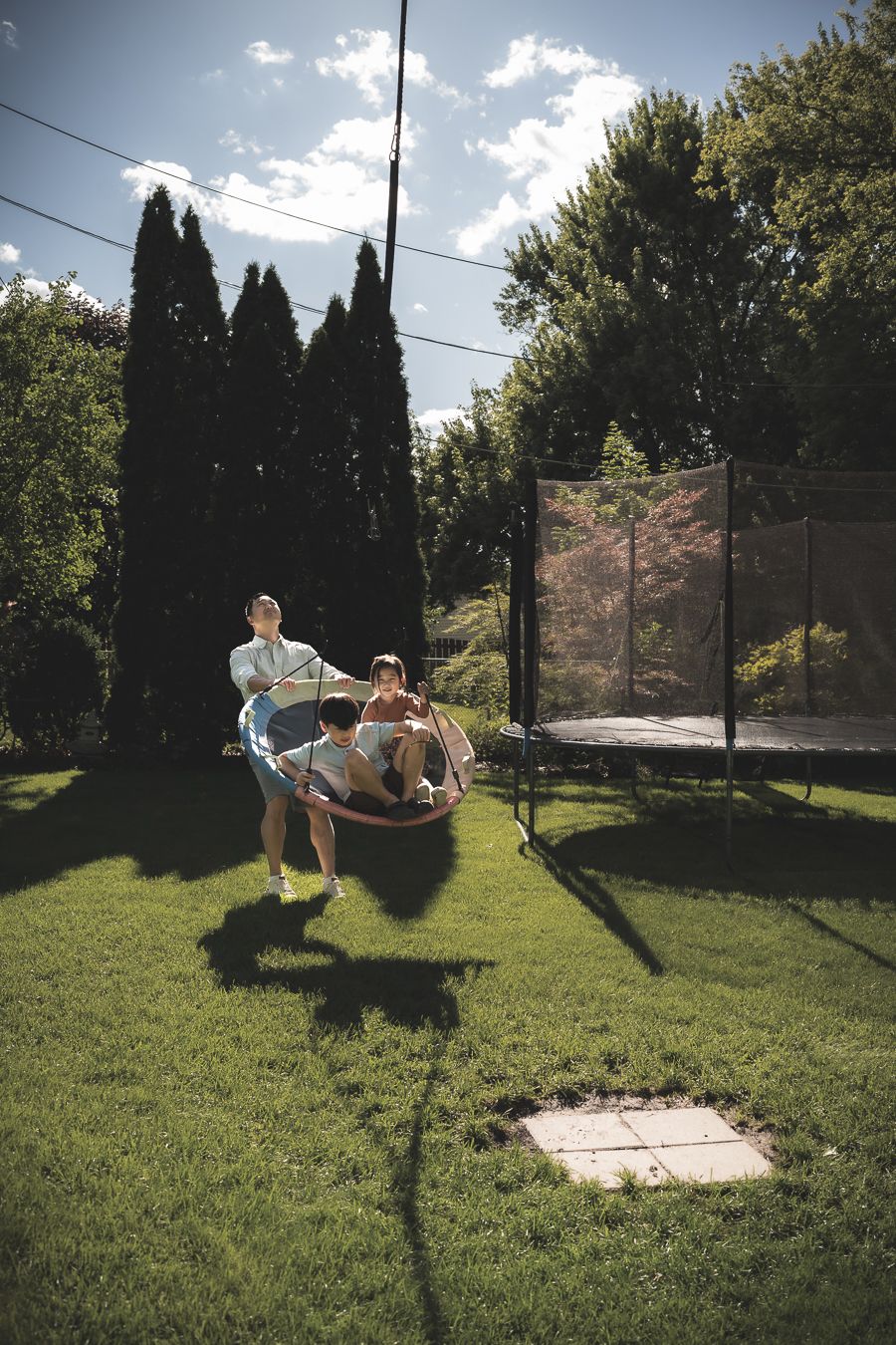 Children playing in a sunny backyard with a large trampoline, one riding in a wheelbarrow on lush green grass.