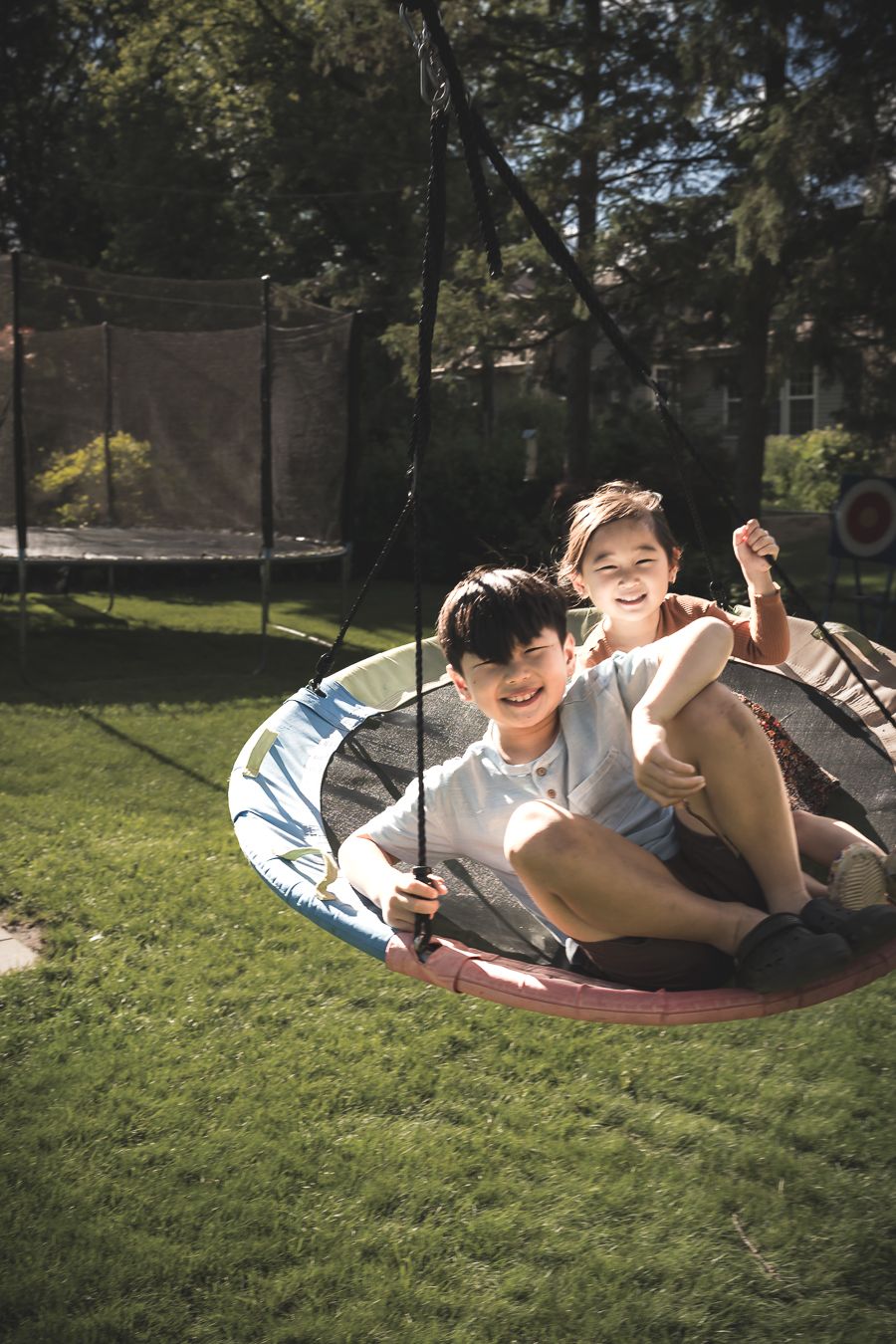 Two happy children laughing and playing on a large round nest swing in a sunny backyard with a trampoline.