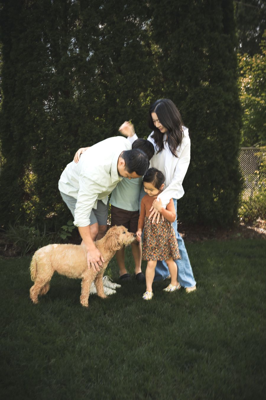 A happy family with two children petting their golden doodle dog together in a lush green backyard.