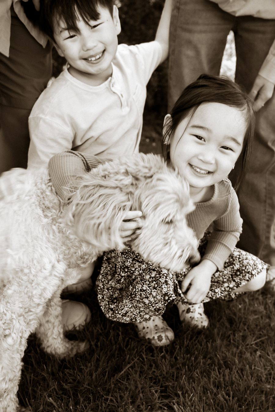 Two smiling children playing with a fluffy white dog outdoors in a sepia-toned family photo.
