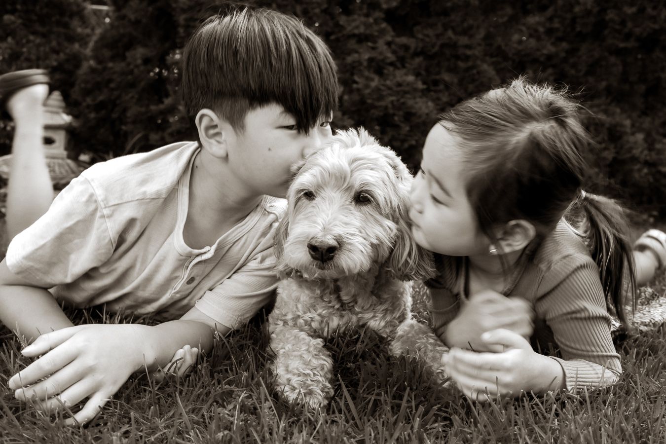 Two children kissing a fluffy puppy on the cheeks while lying on grass in a sweet black and white photo.