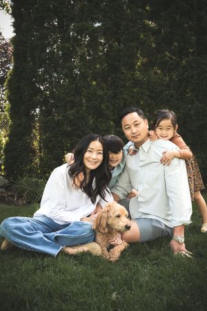 Asian family of four poses outdoors on grass with golden doodle dog, smiling warmly in front of lush green trees.