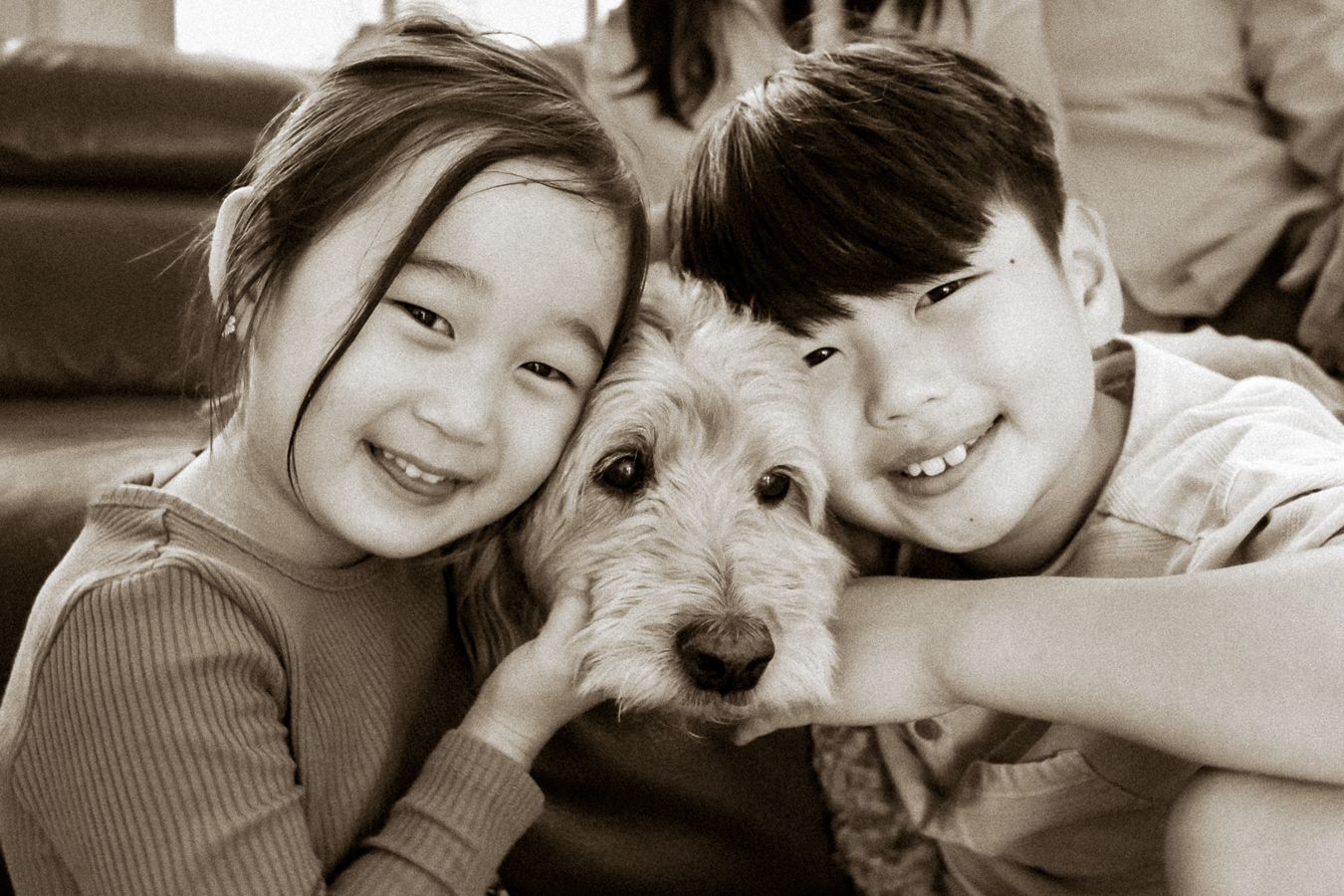 Two smiling children hugging a fluffy terrier dog in a warm sepia-toned portrait photo.