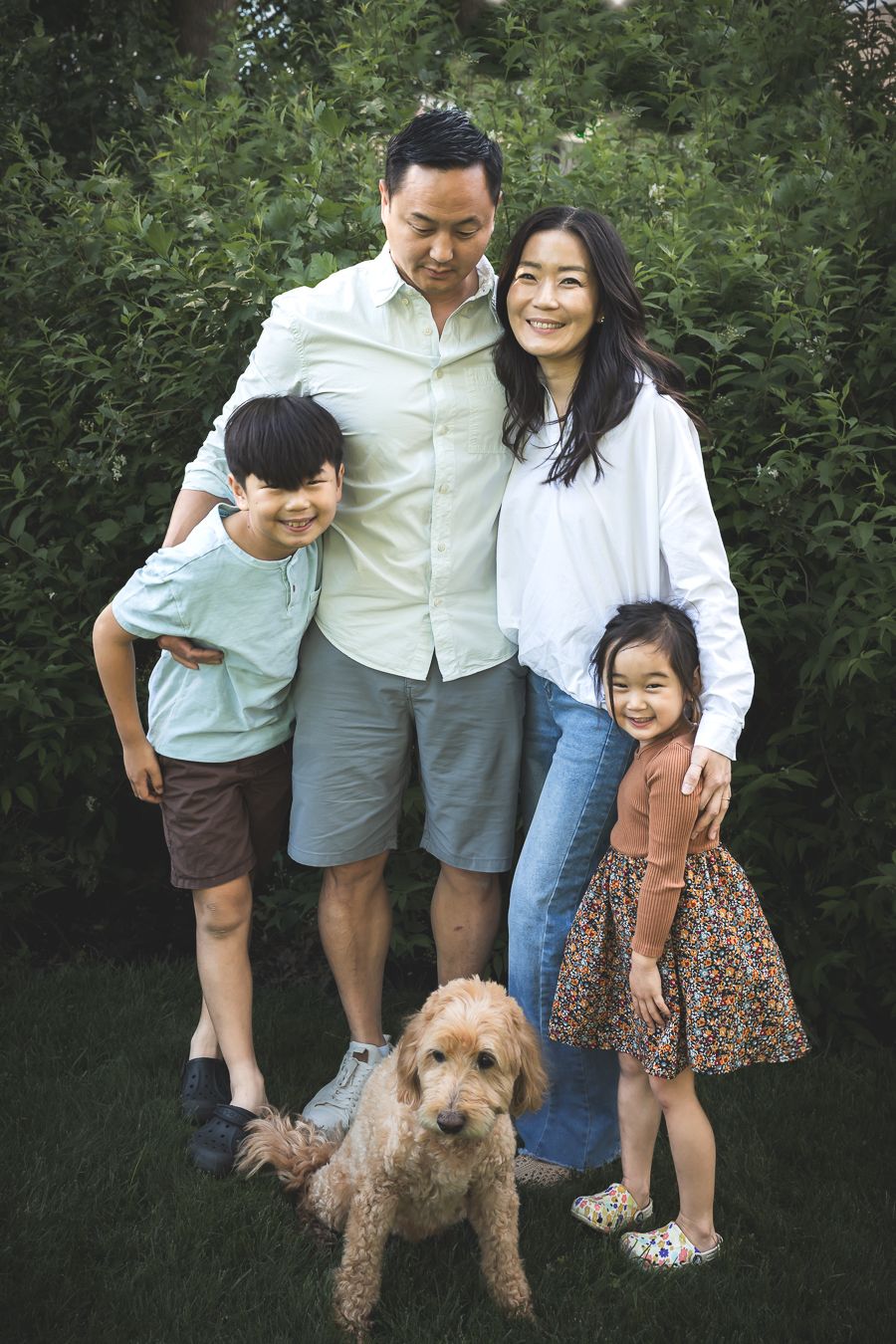 Asian family of four with a golden doodle dog posing outdoors in a lush green garden, smiling together.