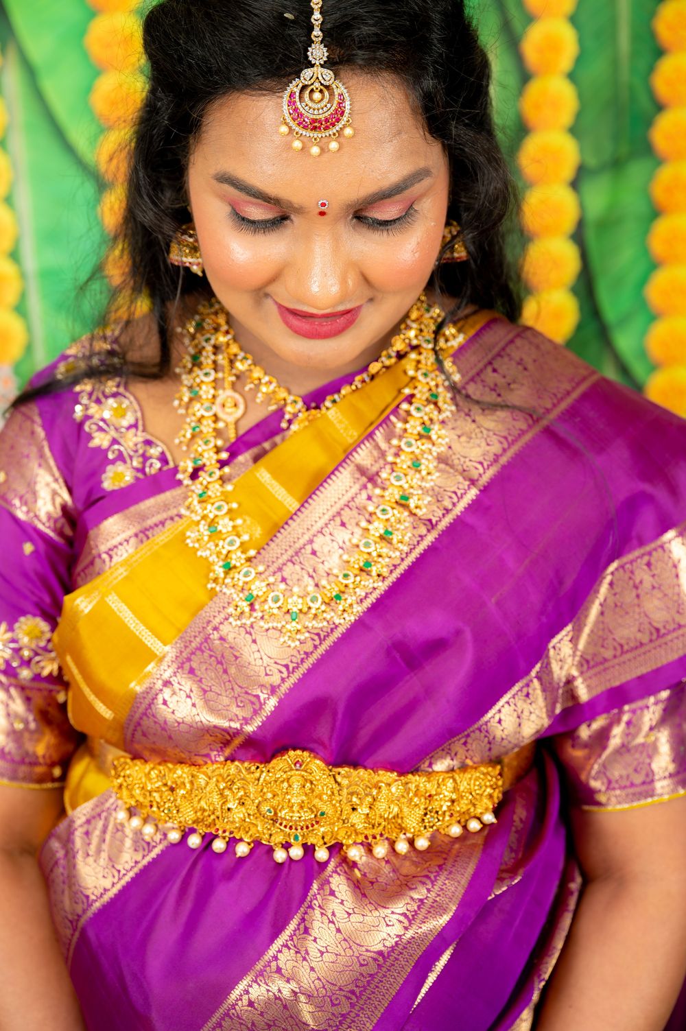 Indian bride in purple and gold silk saree with traditional jewelry, maang tikka, and floral backdrop.