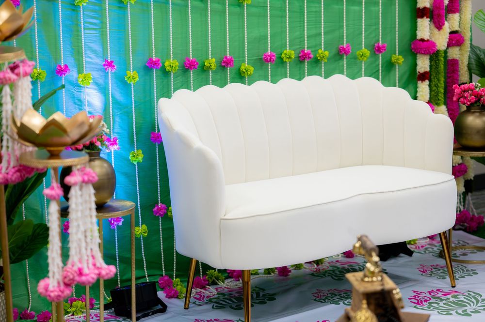White velvet scalloped loveseat sofa with gold legs against a vibrant green floral backdrop at an Indian wedding.