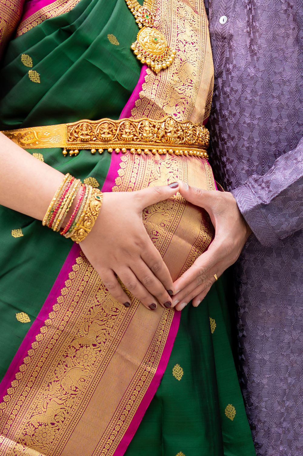 Pregnant woman in green and gold saree with bangles, partner's hands forming heart shape on baby bump.