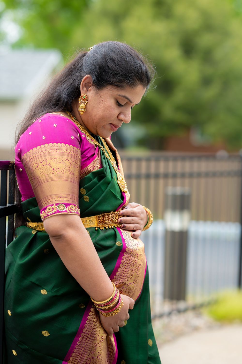 Woman in green and pink silk saree with gold jewelry poses outdoors in traditional South Indian attire.