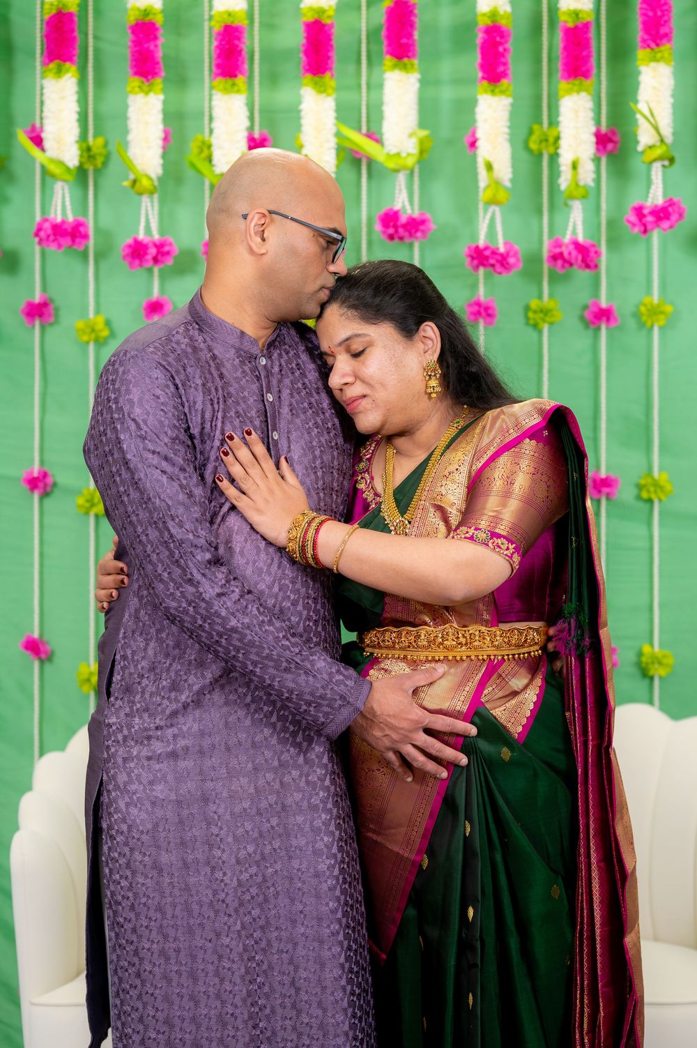Indian couple in traditional attire embracing at a decorated ceremony with floral garlands backdrop.