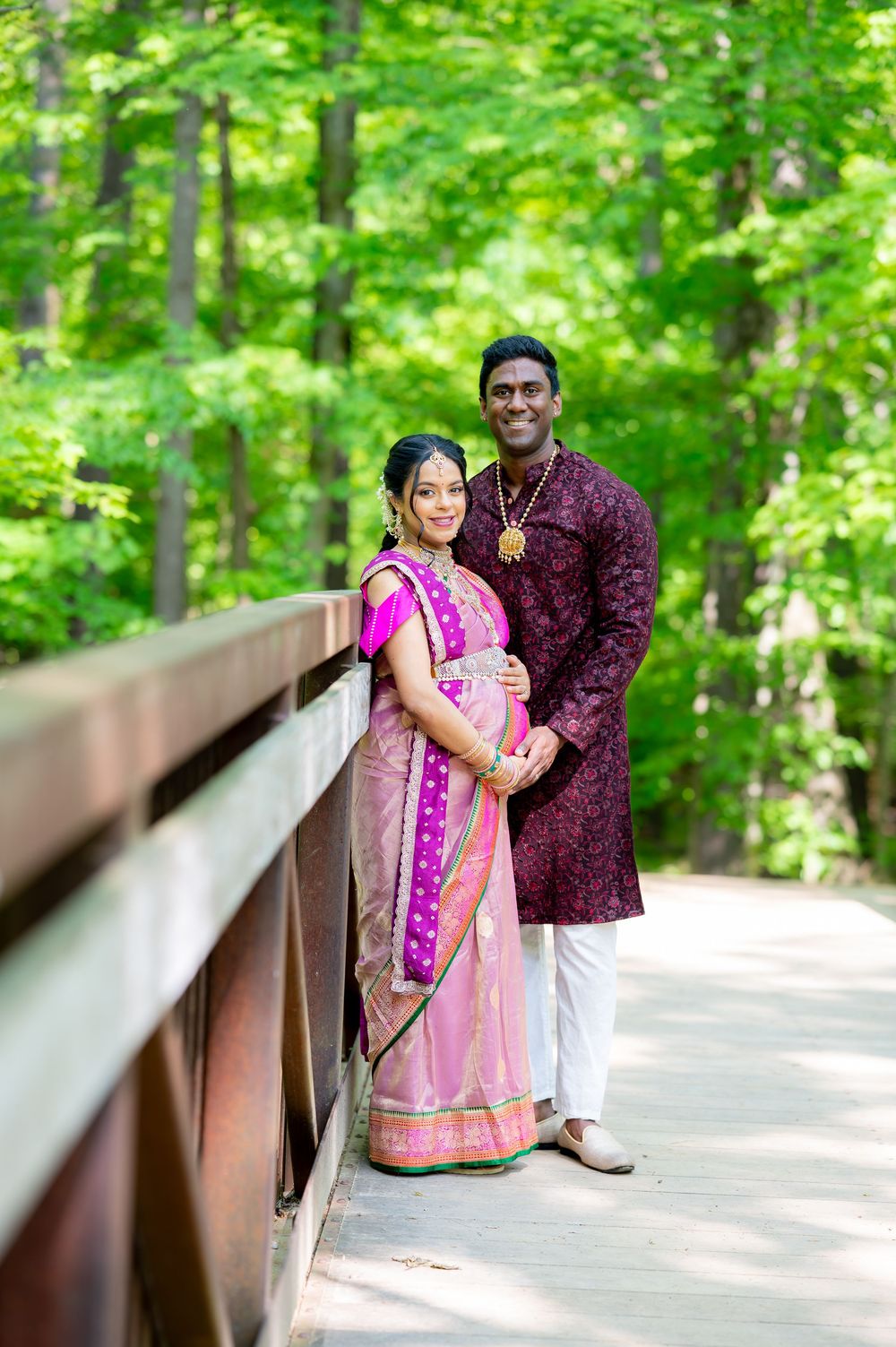 Pregnant couple in traditional Indian attire posing on a wooden bridge surrounded by lush green trees.