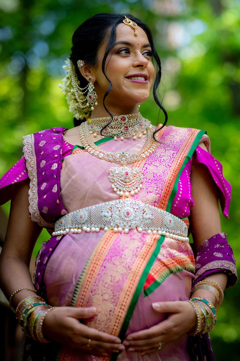 Pregnant Indian bride in pink silk saree with gold jewelry and floral accessories during a traditional baby shower ceremony.