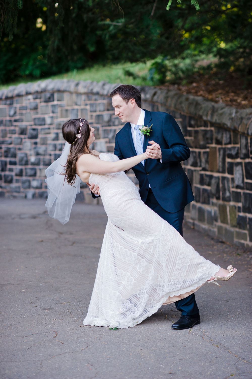 Groom in navy suit dipping bride in fitted lace wedding dress near rustic stone wall outdoors.