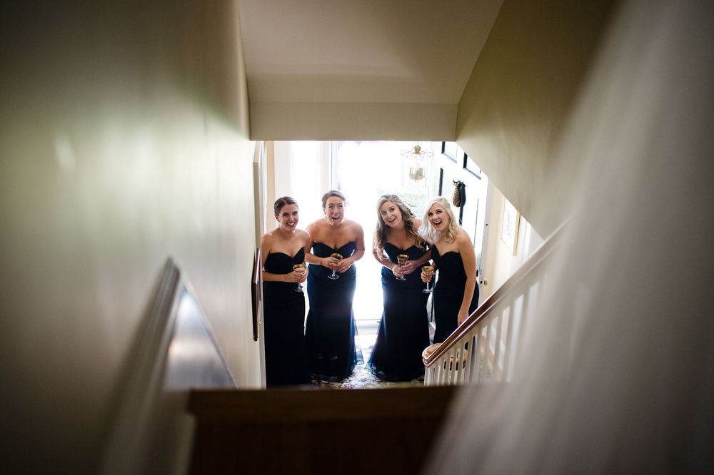 Four bridesmaids in navy blue strapless gowns stand at the bottom of a staircase, smiling up toward the camera.