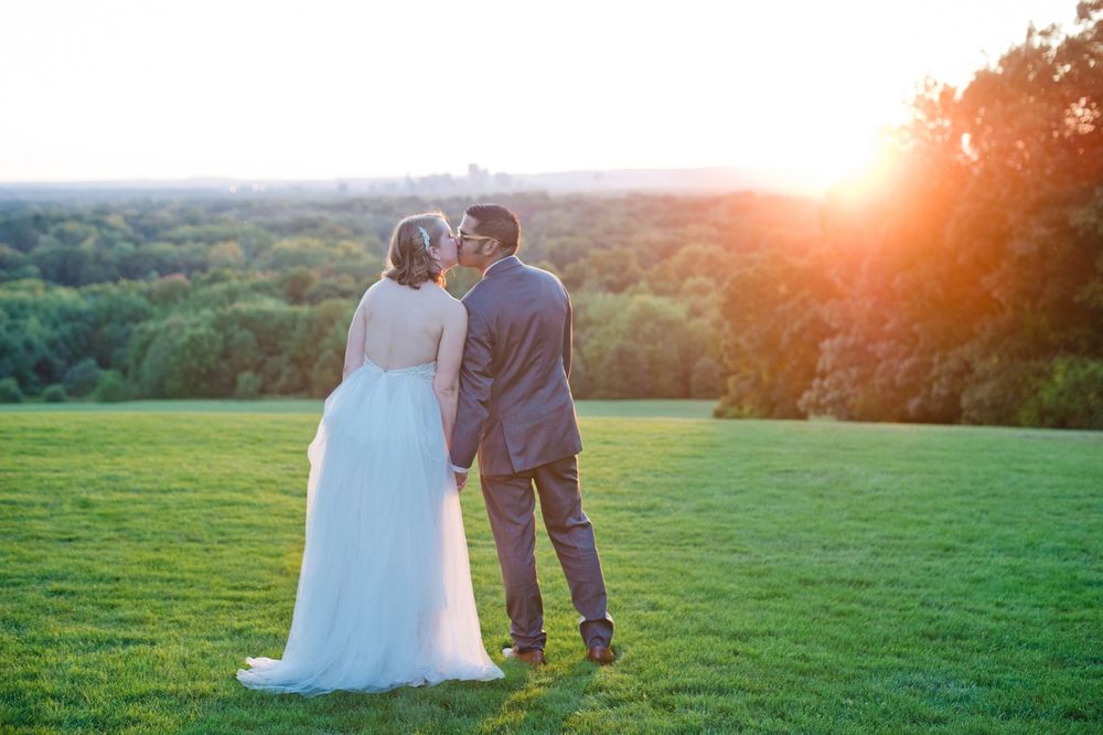 Bride and groom sharing a romantic sunset kiss on a green hillside with city skyline in background.
