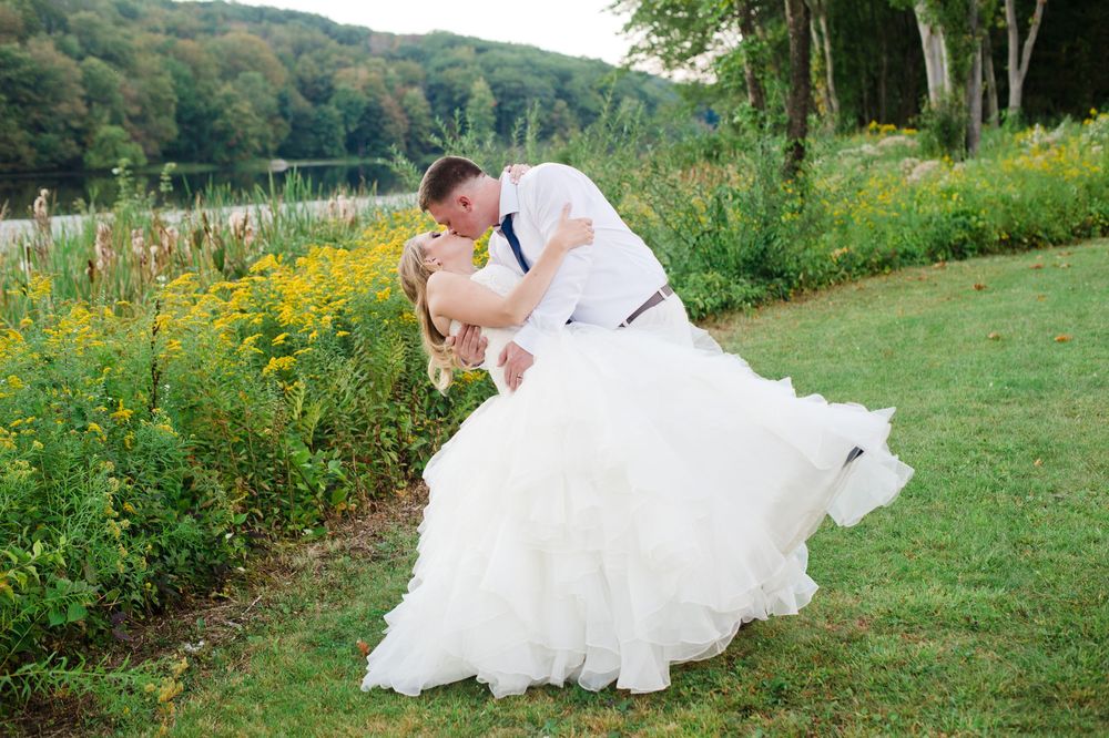 Groom dipping bride in layered white gown for a romantic kiss among wildflowers by a serene lake.