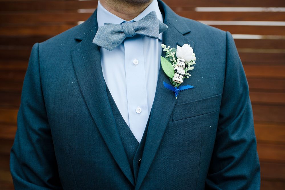 Groom wearing a teal three-piece suit with a grey bow tie and a white floral boutonniere with greenery accents.