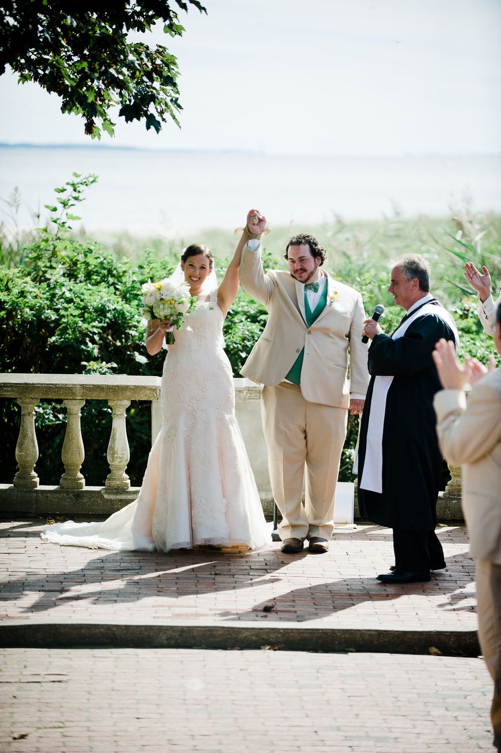 Bride and groom raise hands in celebration after outdoor wedding ceremony near waterfront, officiant applauds beside them.