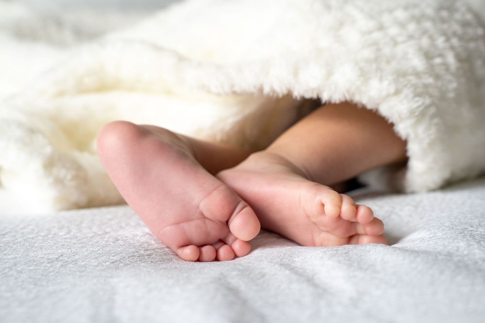 Tiny newborn baby feet peeking out from under a soft white fluffy blanket on a white bed.