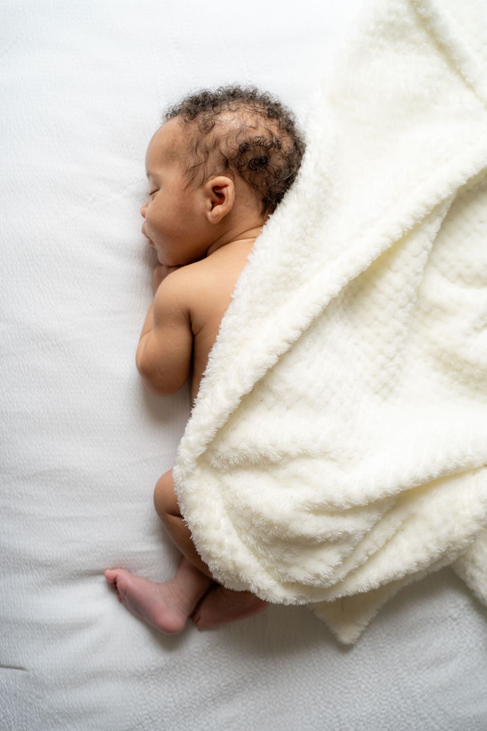 Newborn baby sleeping peacefully on white bed, partially covered with soft cream blanket, viewed from above.