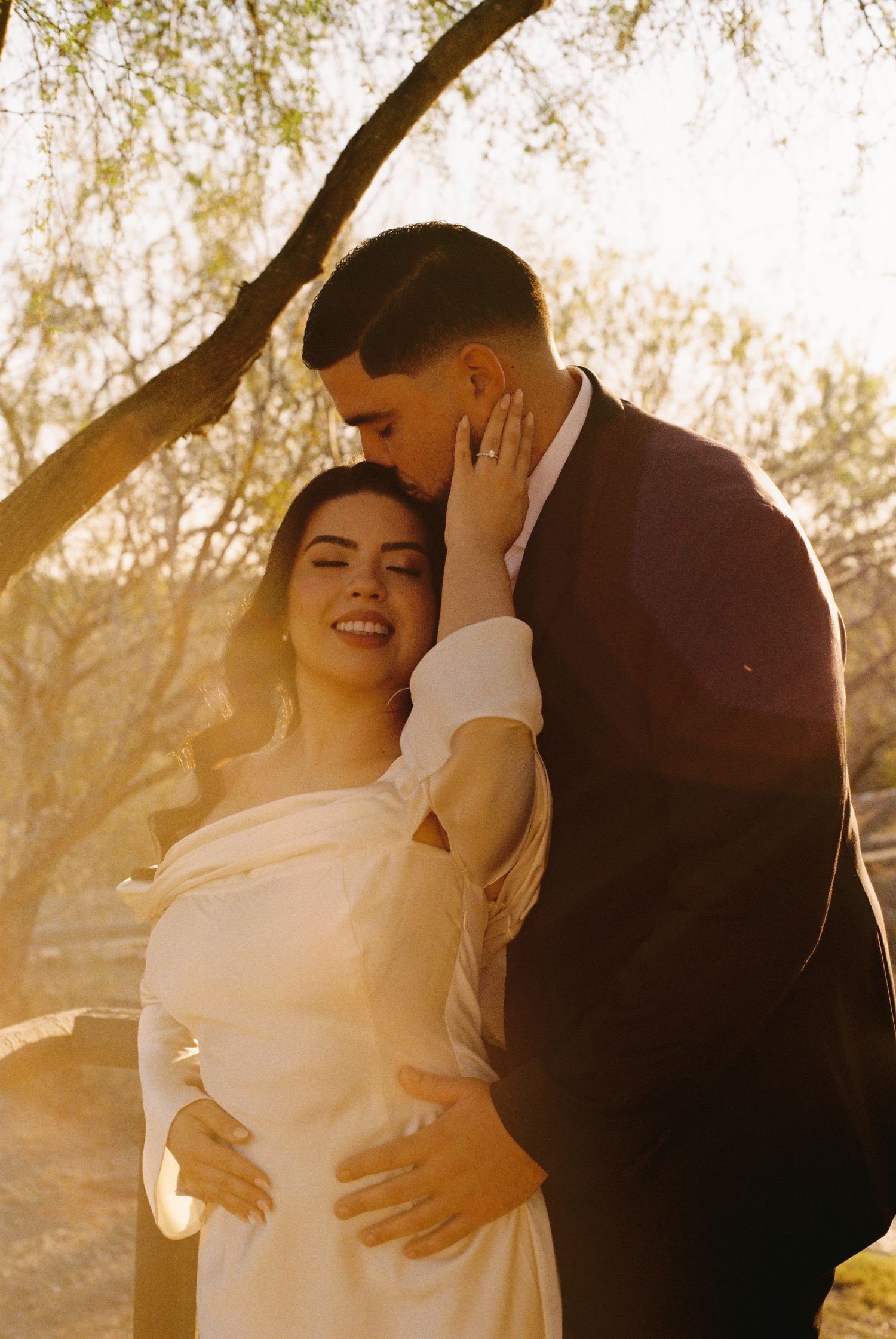 A bride in a white gown smiles as her groom in a dark suit kisses her head during a golden hour outdoor wedding.