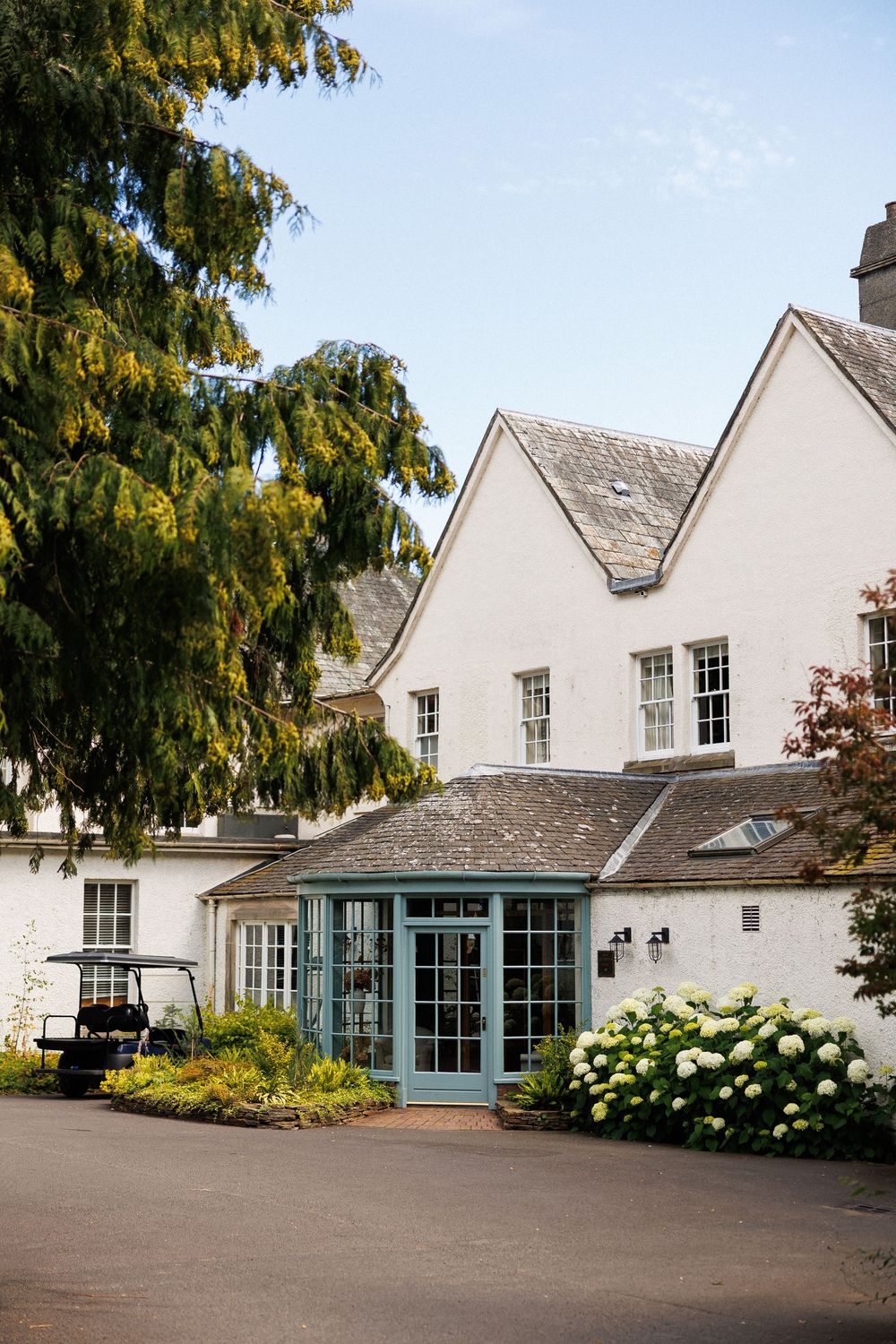 White country house hotel with teal glass entrance, hydrangeas, and mature trees under a clear blue sky.