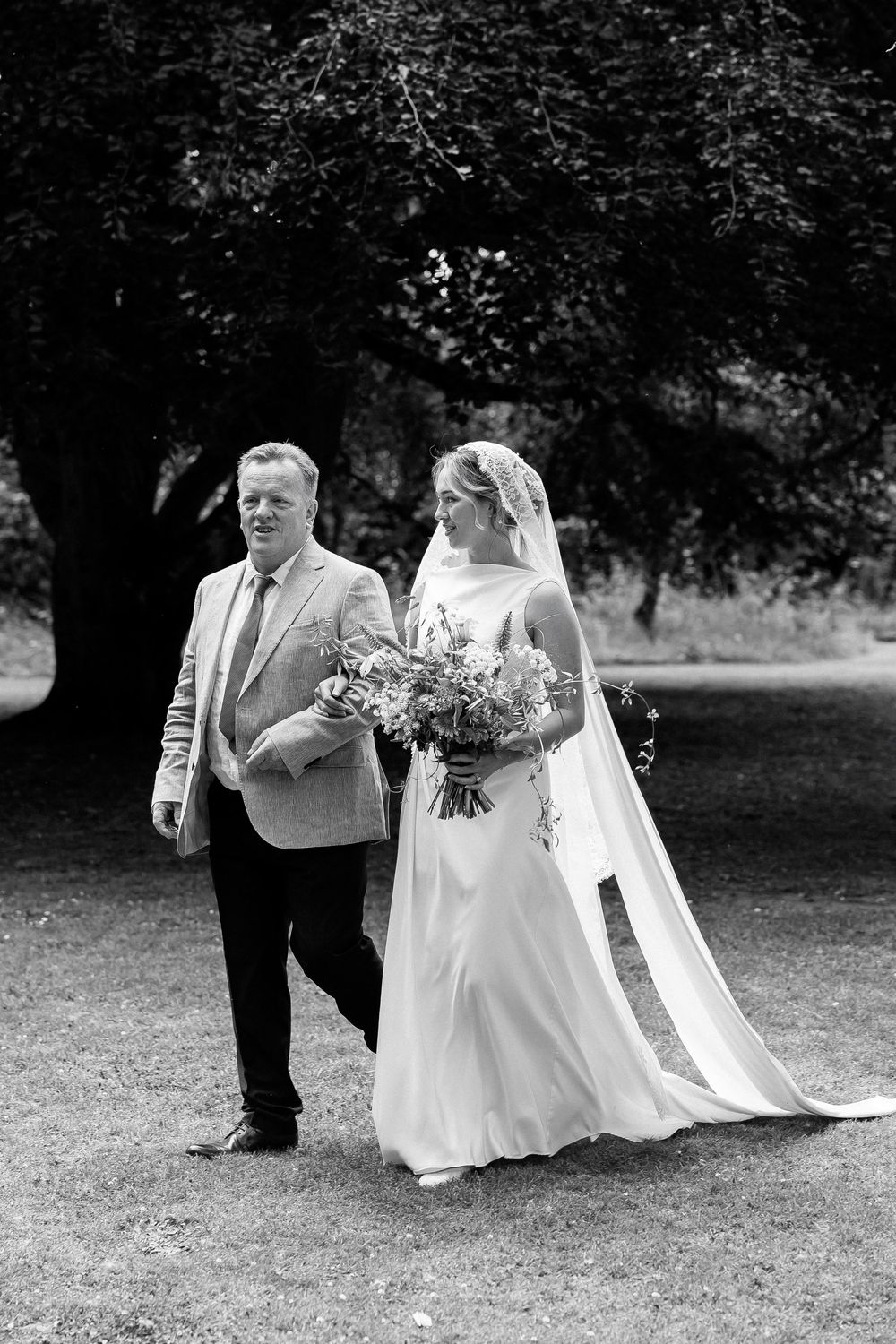 Black and white photo of a bride holding a bouquet walking with a man in a suit through a garden.