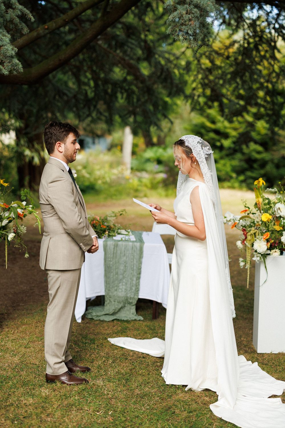 Bride reads vows to groom during outdoor garden wedding ceremony with floral arrangements and lush green trees.