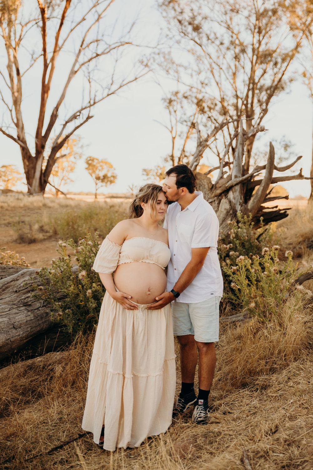 Couple sharing a kiss in rustic Australian bushland, showcasing pregnant belly at golden hour.
