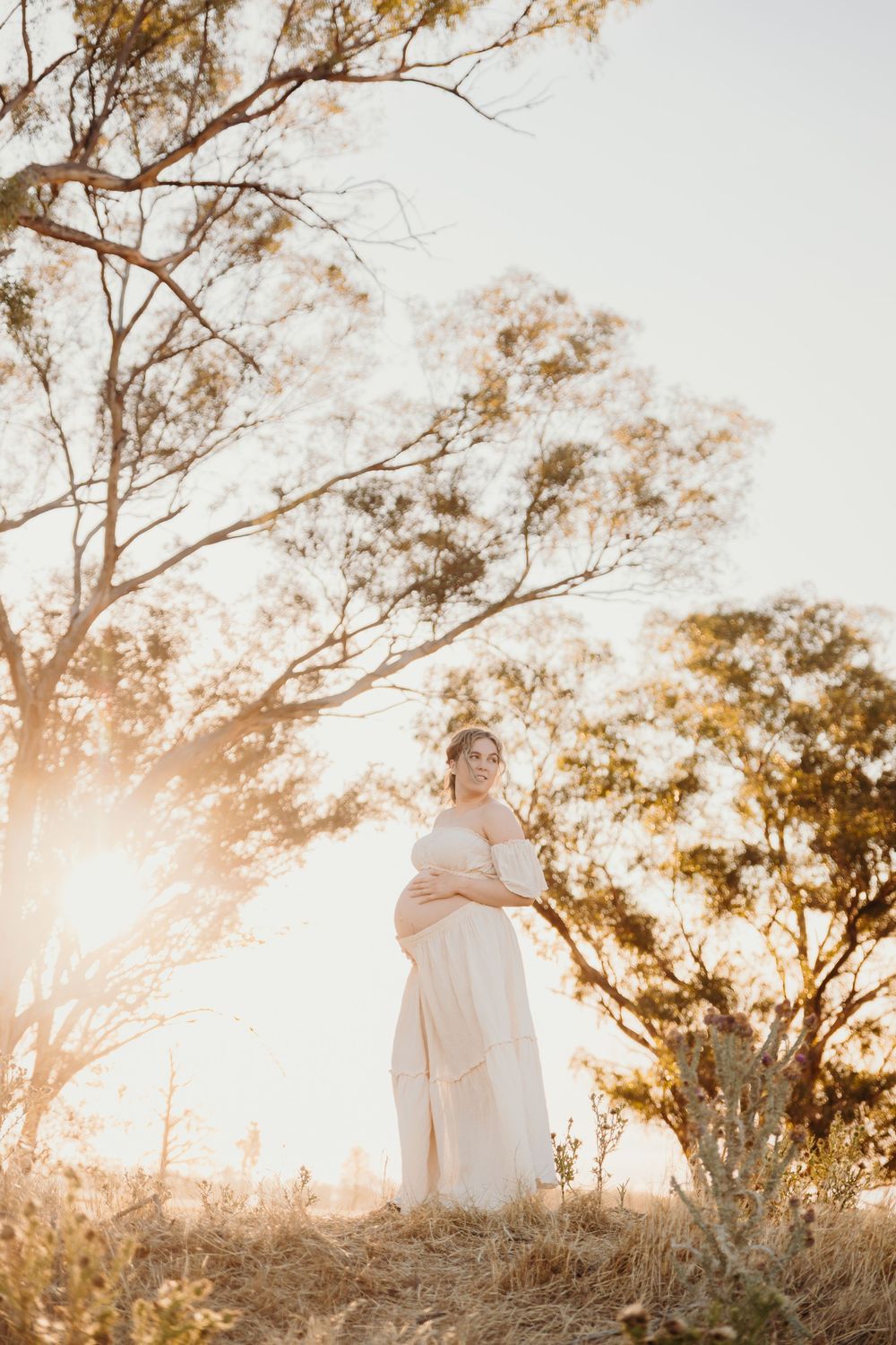 Glowing pregnant woman in cream dress standing under eucalyptus trees in golden sunset light.