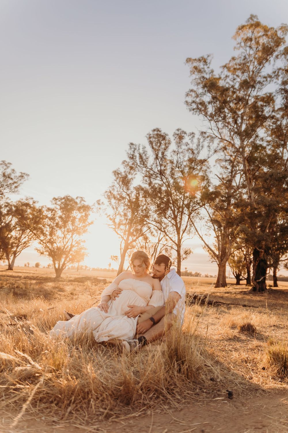 A pregnant couple sits together in a golden field at sunset surrounded by eucalyptus trees.