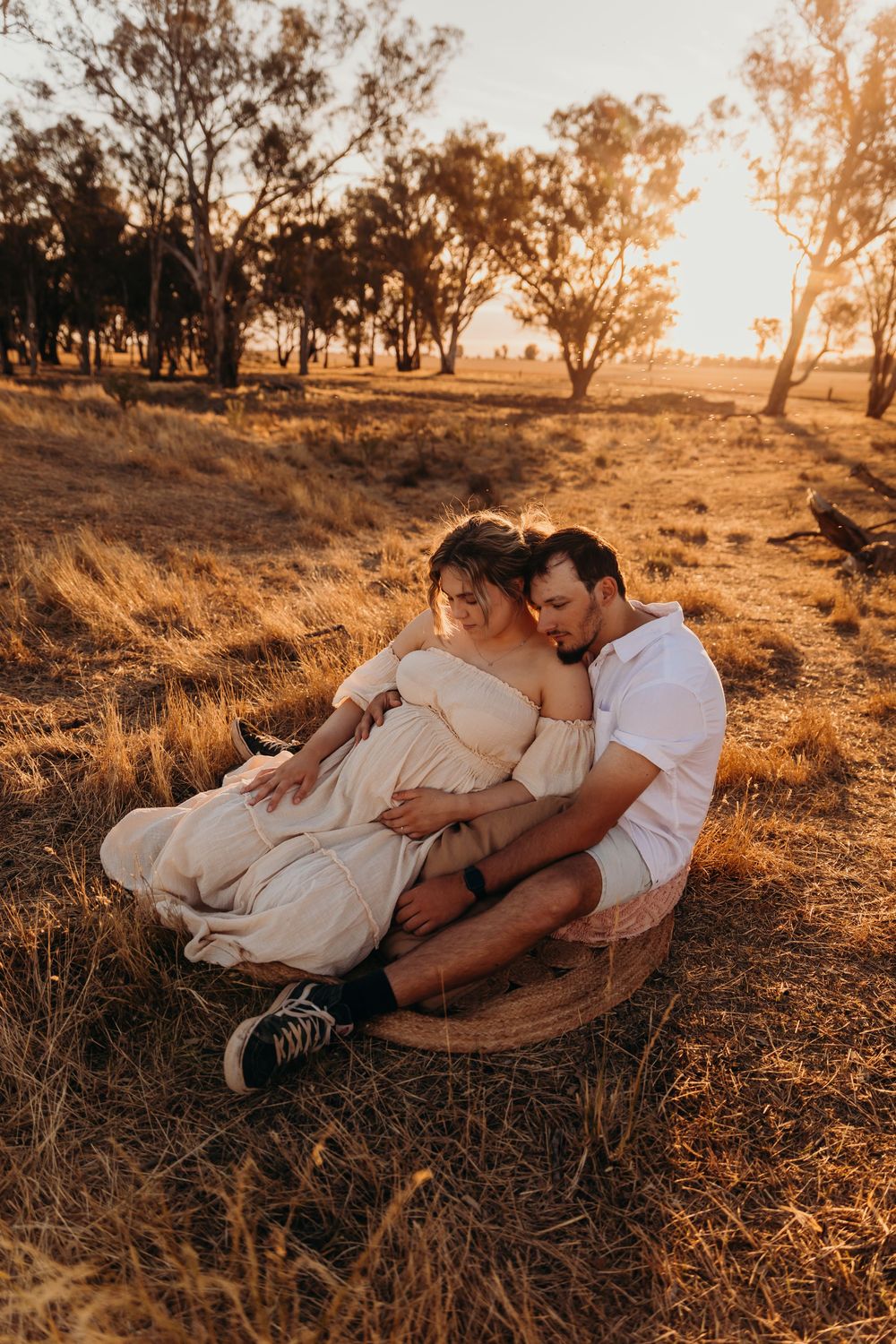 A couple embraces in a dry golden field at sunset, the woman visibly pregnant.