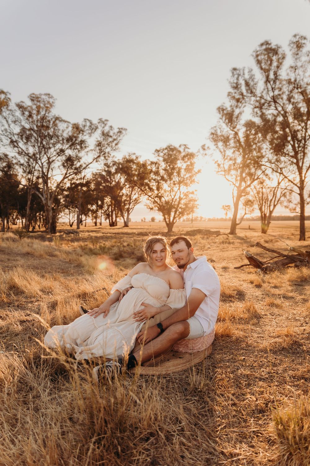 A pregnant woman in a cream dress sits with her partner in a sunlit golden field.