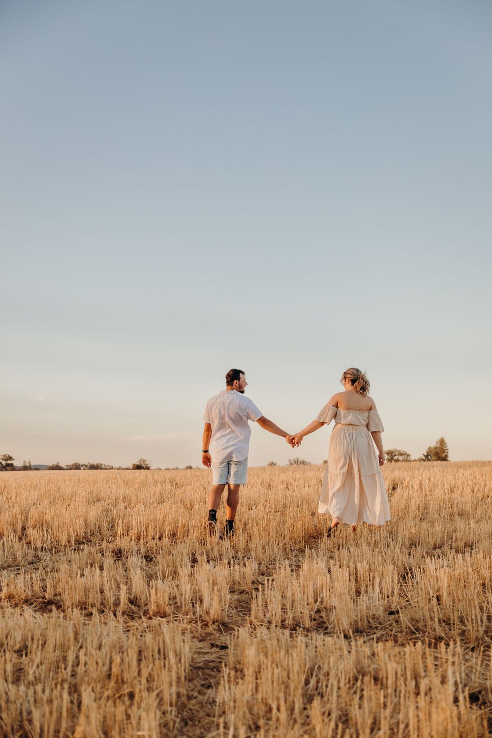 A couple holds hands walking through a golden wheat field at sunset.
