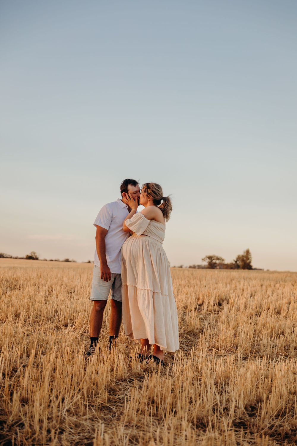 A couple shares a kiss in a vast golden wheat field during a warm sunset.