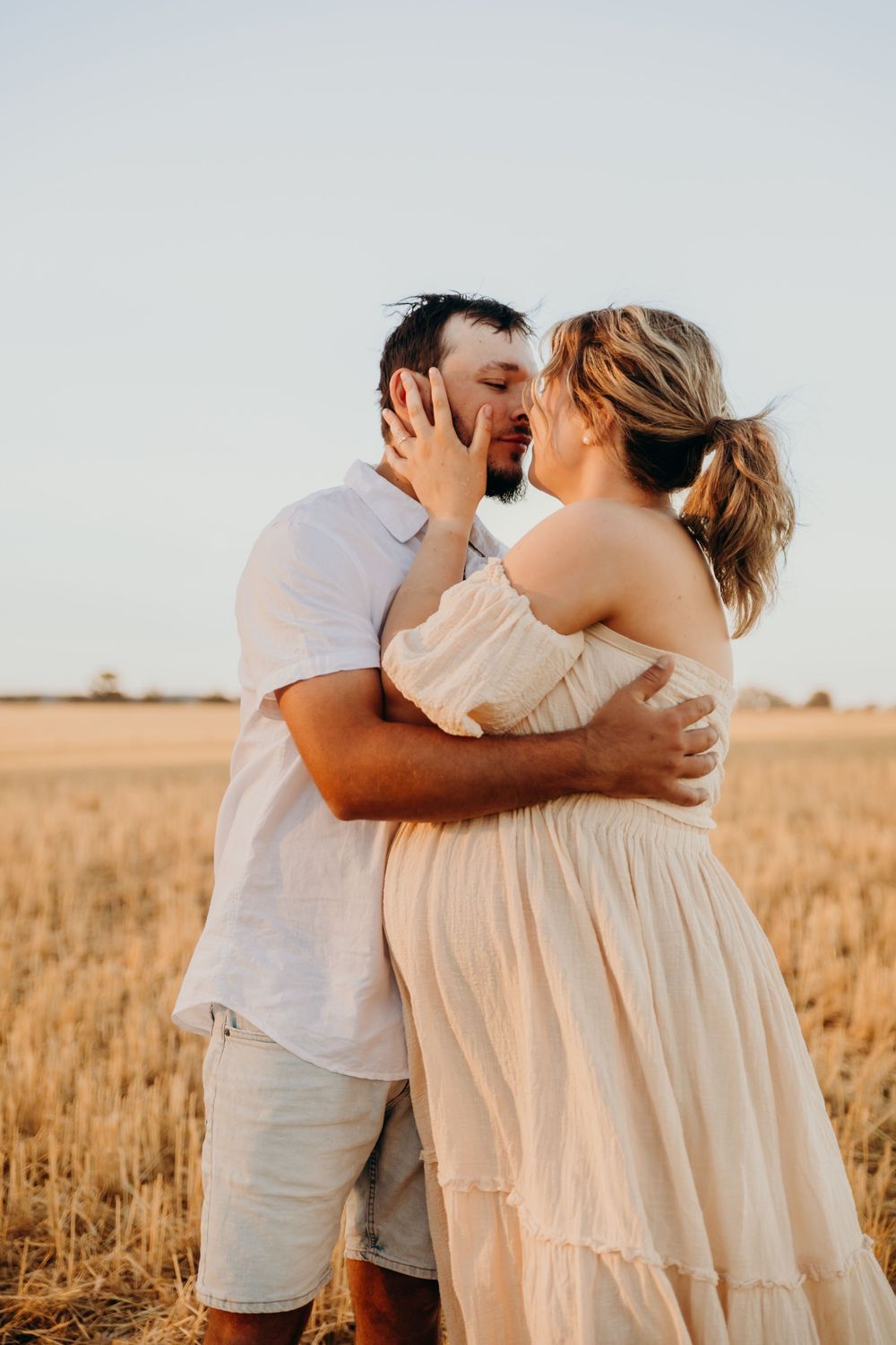 A couple kisses and embraces in a golden wheat field under a clear evening sky.