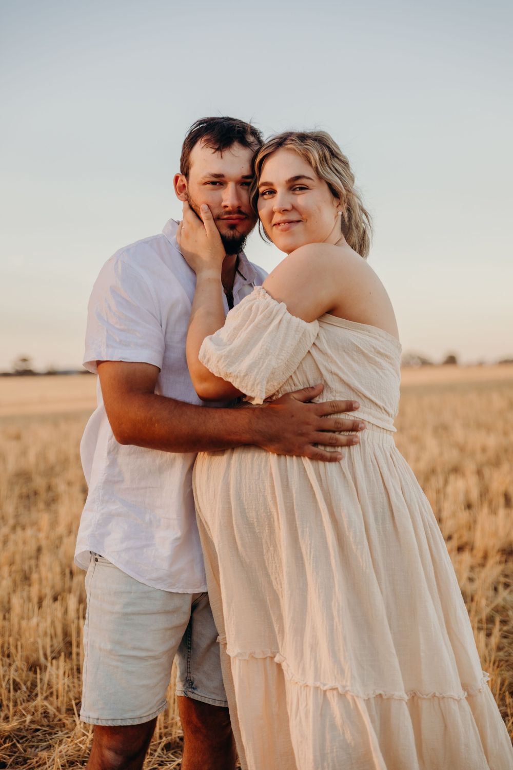 Couple embracing in golden wheat field at sunset, pregnant woman in off-shoulder cream dress.
