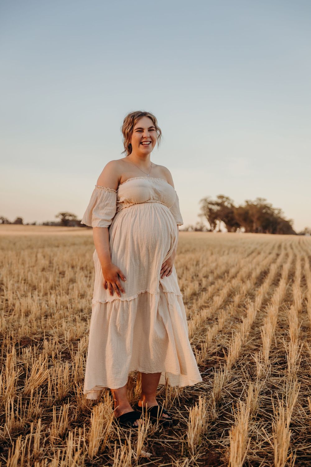 Smiling pregnant woman in cream off-shoulder dress standing in harvested wheat field at sunset.