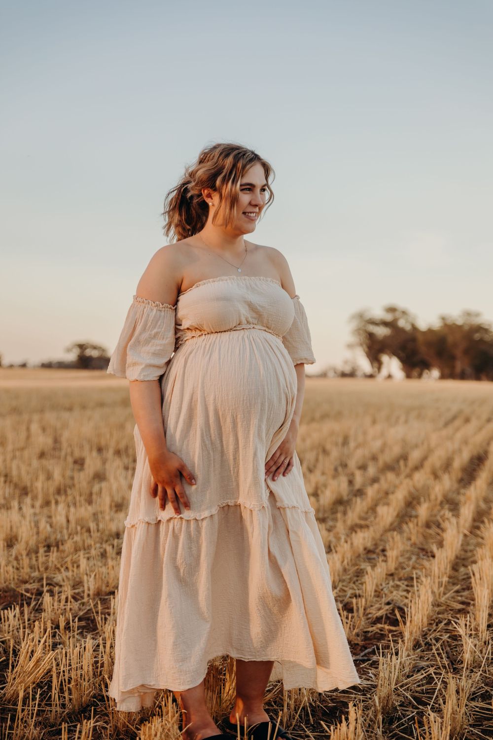 Glowing pregnant woman in flowing cream dress laughing in golden wheat field at sunset.
