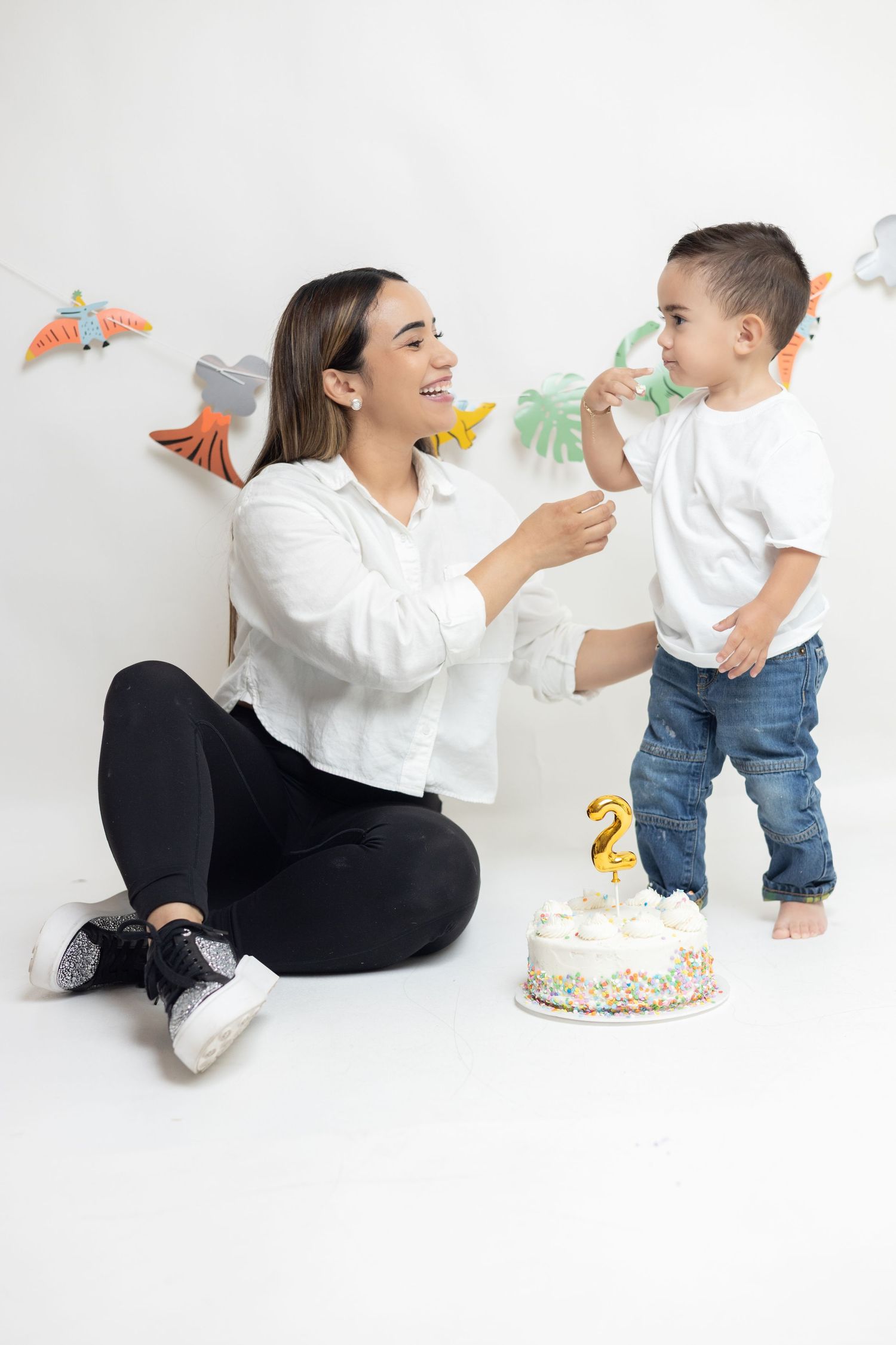 A woman sits on the floor smiling at a toddler boy standing beside a birthday cake with a number 2 candle.