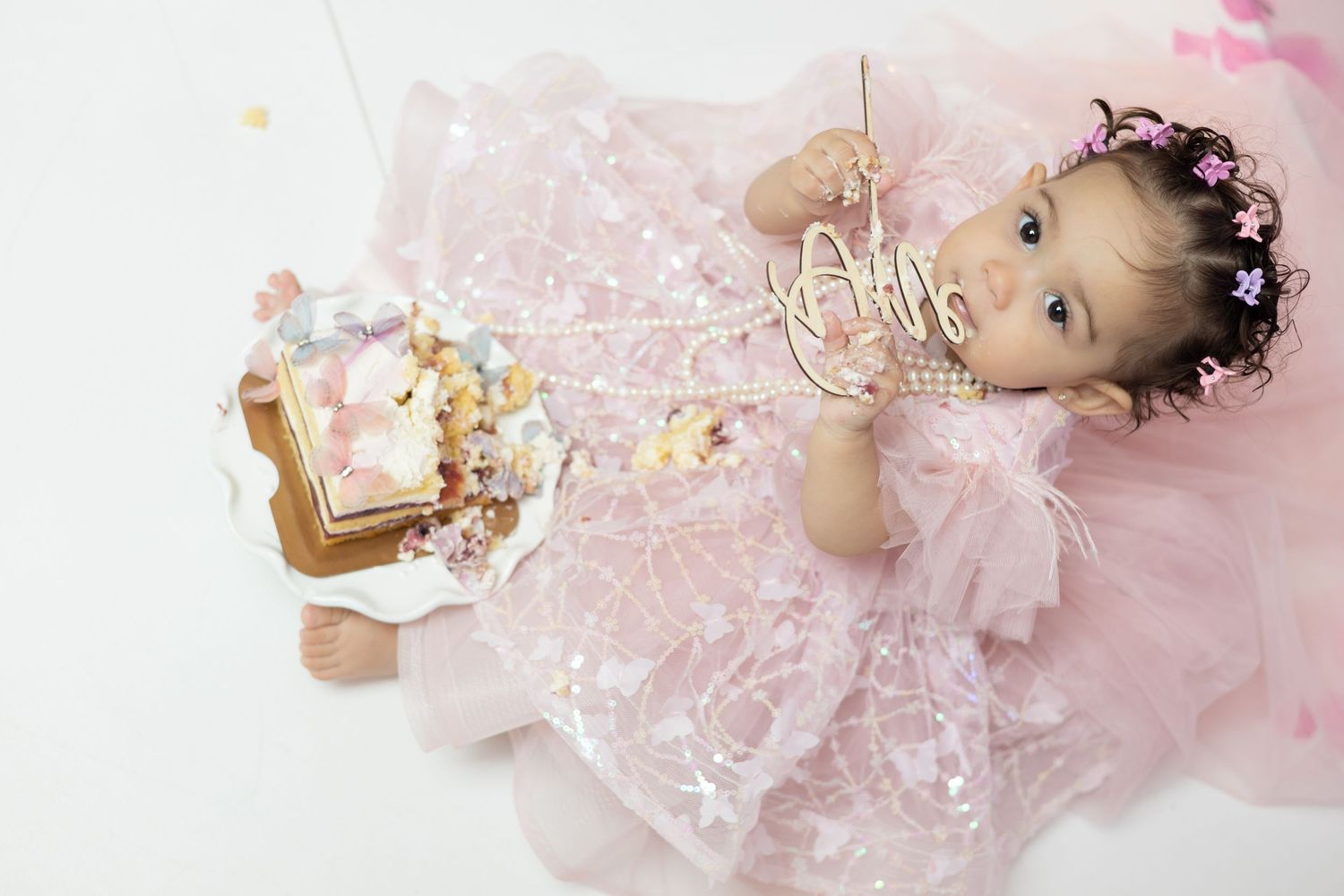 Baby girl in pink tutu dress with floral crown celebrates first birthday with cake smash on white background.