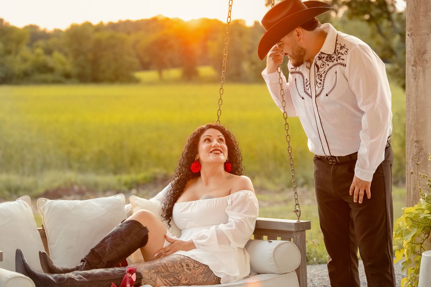 Couple in western attire at golden hour, woman in white off-shoulder dress on swing, man in cowboy hat smiling.