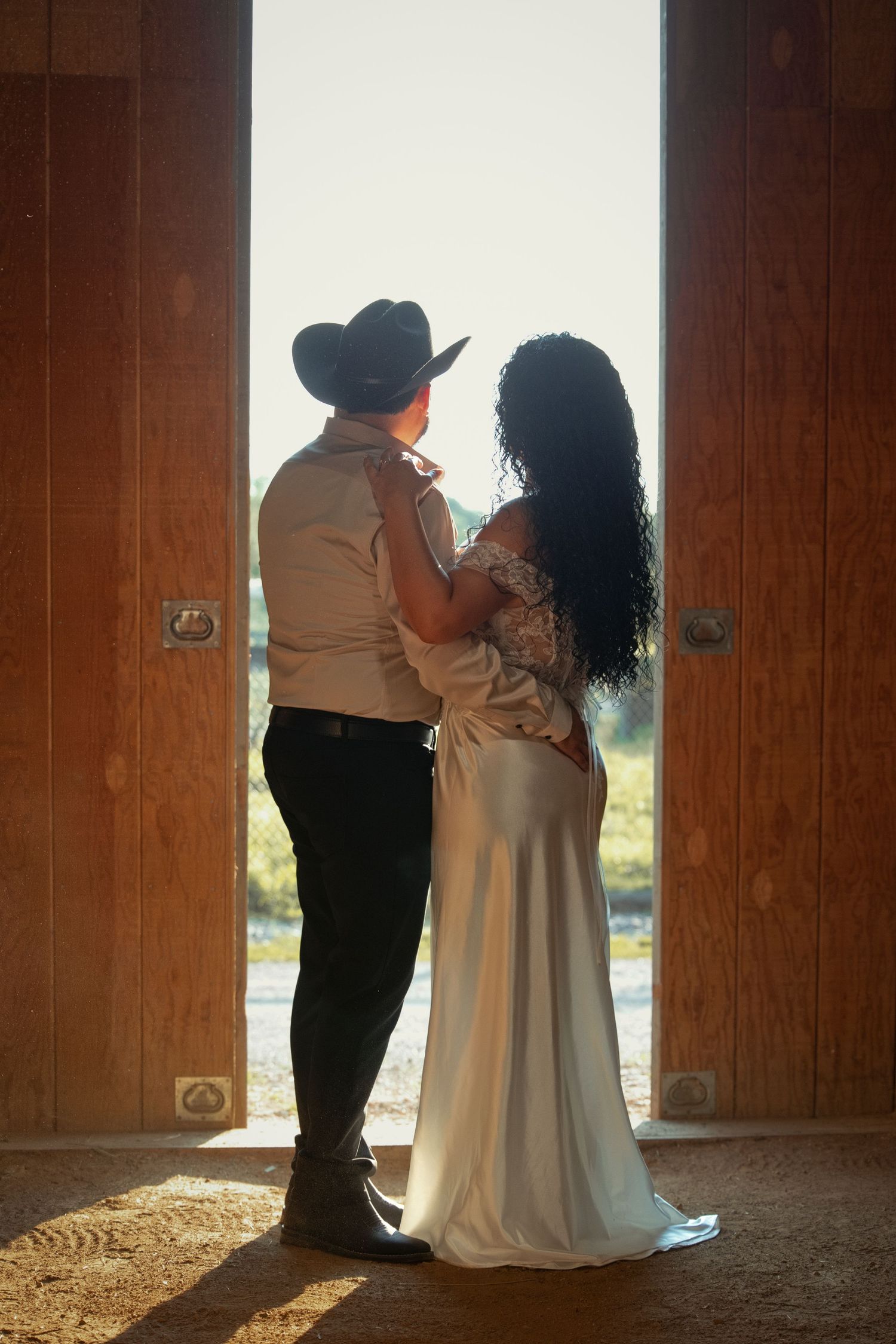Couple embracing in barn doorway, groom in cowboy hat and bride in white gown, backlit by golden sunlight.