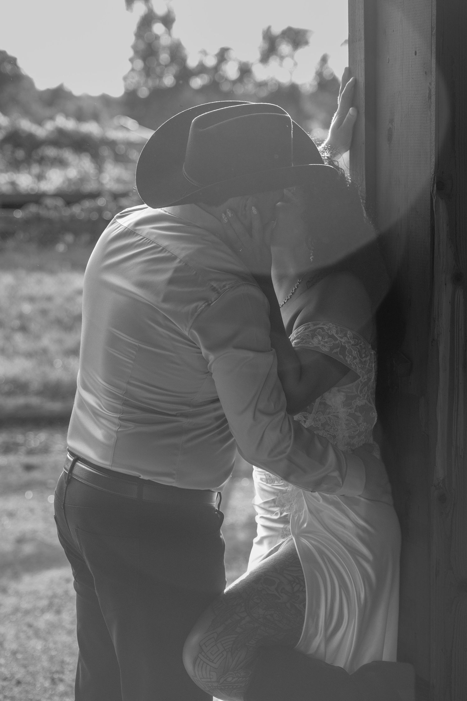 Black and white photo of a couple sharing a romantic kiss, man in cowboy hat and woman in white dress leaning against wooden post.