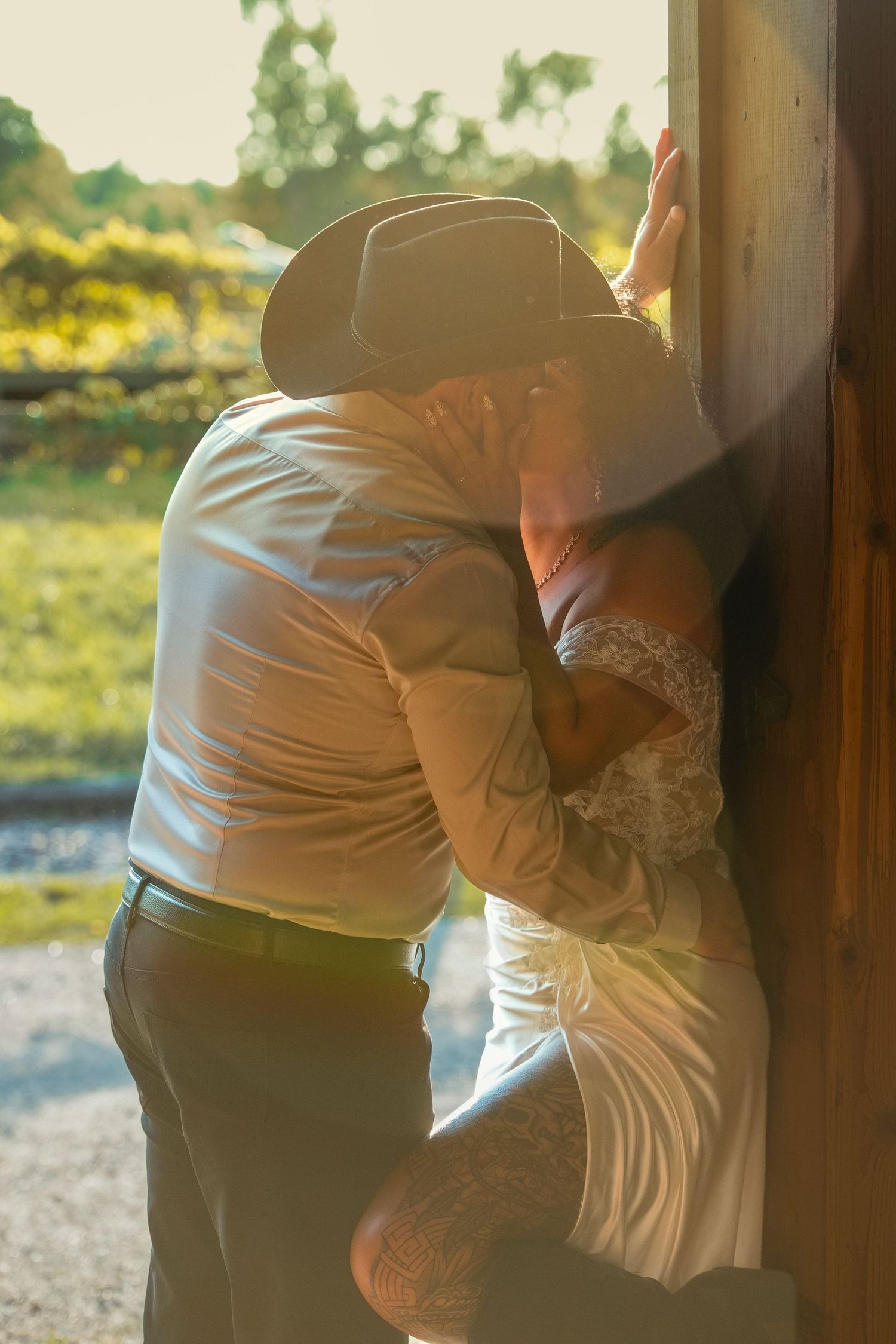 Couple sharing a romantic kiss against a rustic wooden barn post in golden sunset light, groom wearing cowboy hat.