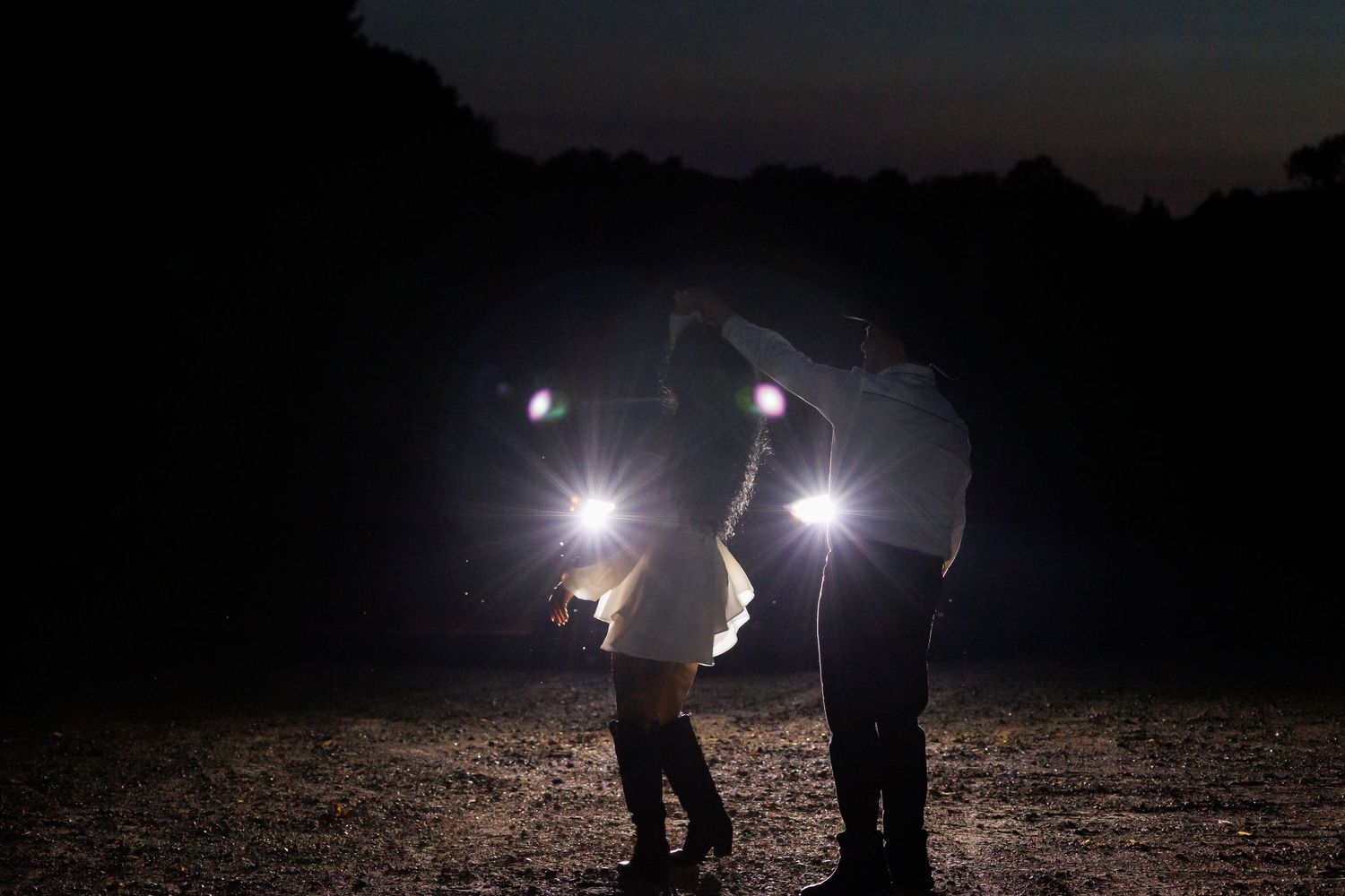 Couple sharing a romantic moment at night, illuminated by bright lights in a dark outdoor setting.