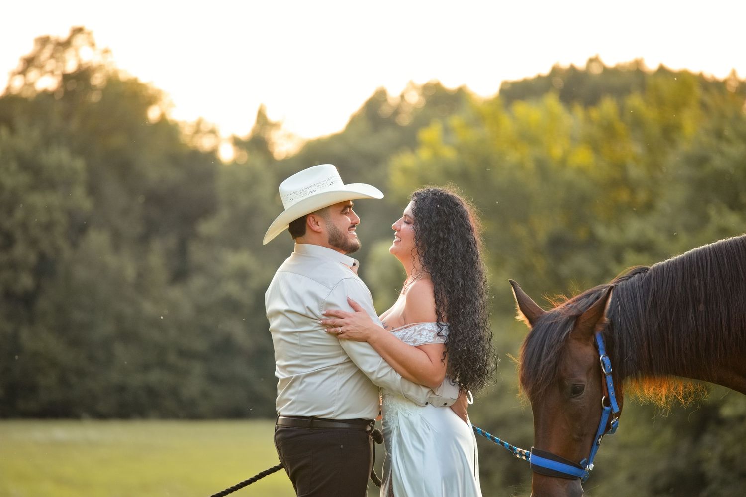 Couple embracing near a brown horse at golden hour in a field, man in cowboy hat, woman in white dress.