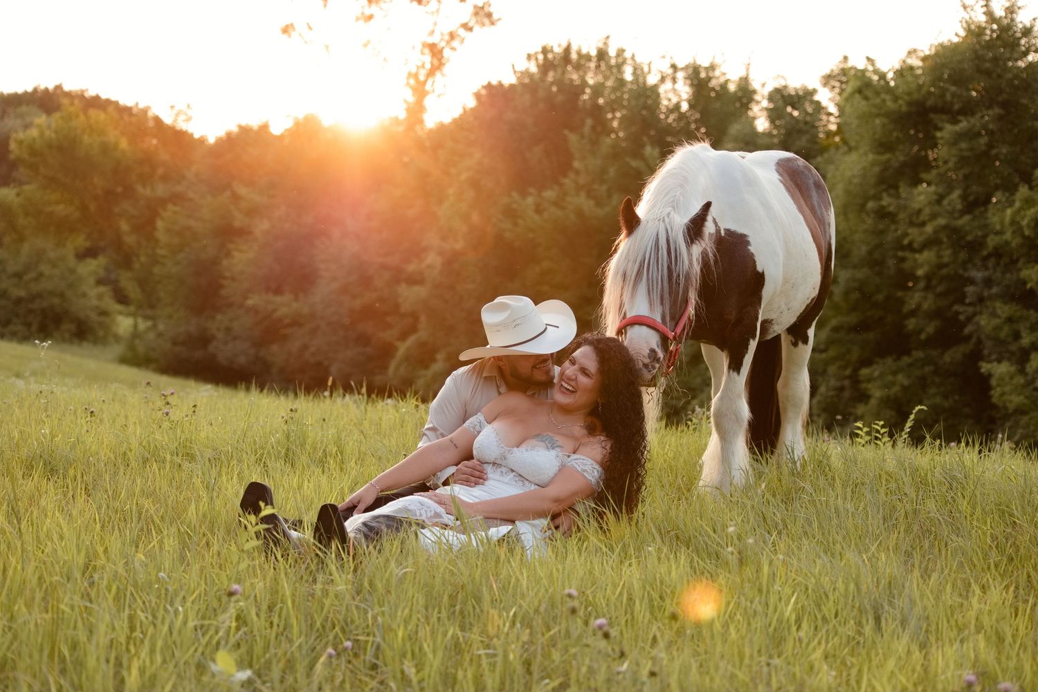 Two women in white dresses sit together in a sunny field beside a paint horse at golden hour.