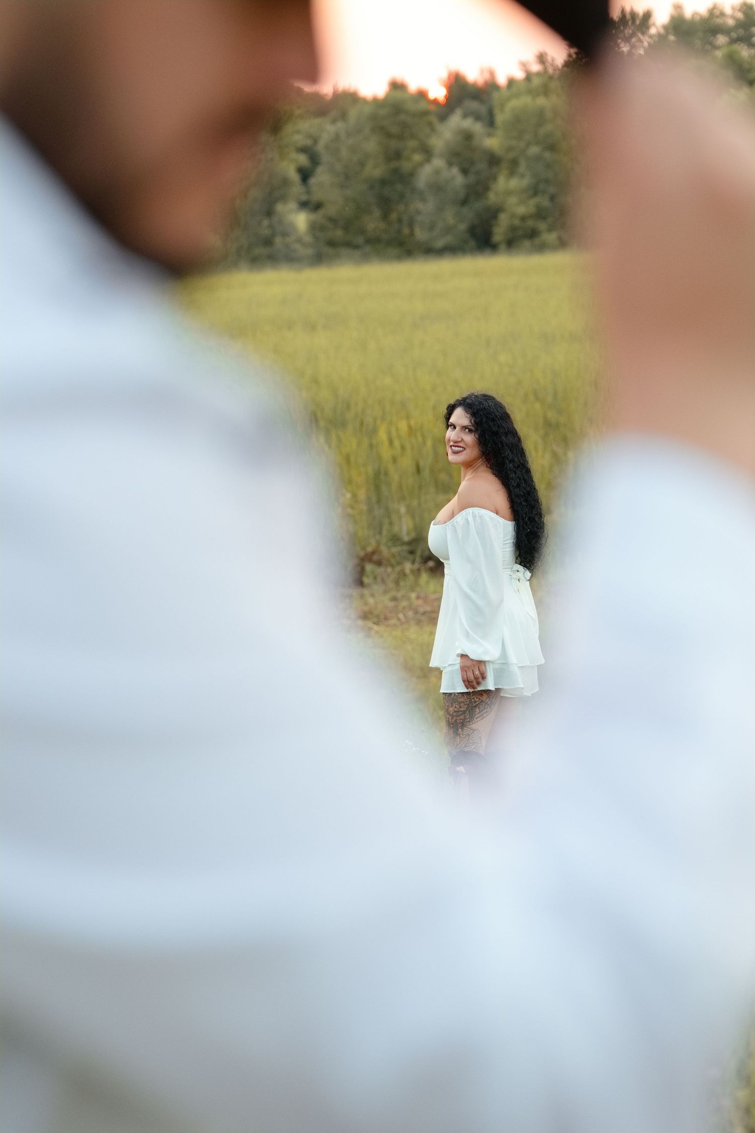 A smiling woman in a white off-shoulder dress stands in a green field, viewed through a blurred foreground figure.